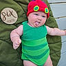 baby, child, infant, green_outfit, red_hat, tongue_out, cushion, plush, wooden_plaque, six_months, outdoor, person, cute, smiling, playful, skin, face, headwear, happy, portrait