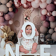 Orla is registered to the contest to win money with this photo: baby, child, one_year_old, birthday, balloons, decorations, letters, white_chair, playful, tongue_out, pastel_colors, boho_decor, round_rug, indoor, portrait, celebration, cute, small_feet, smiling, sitting