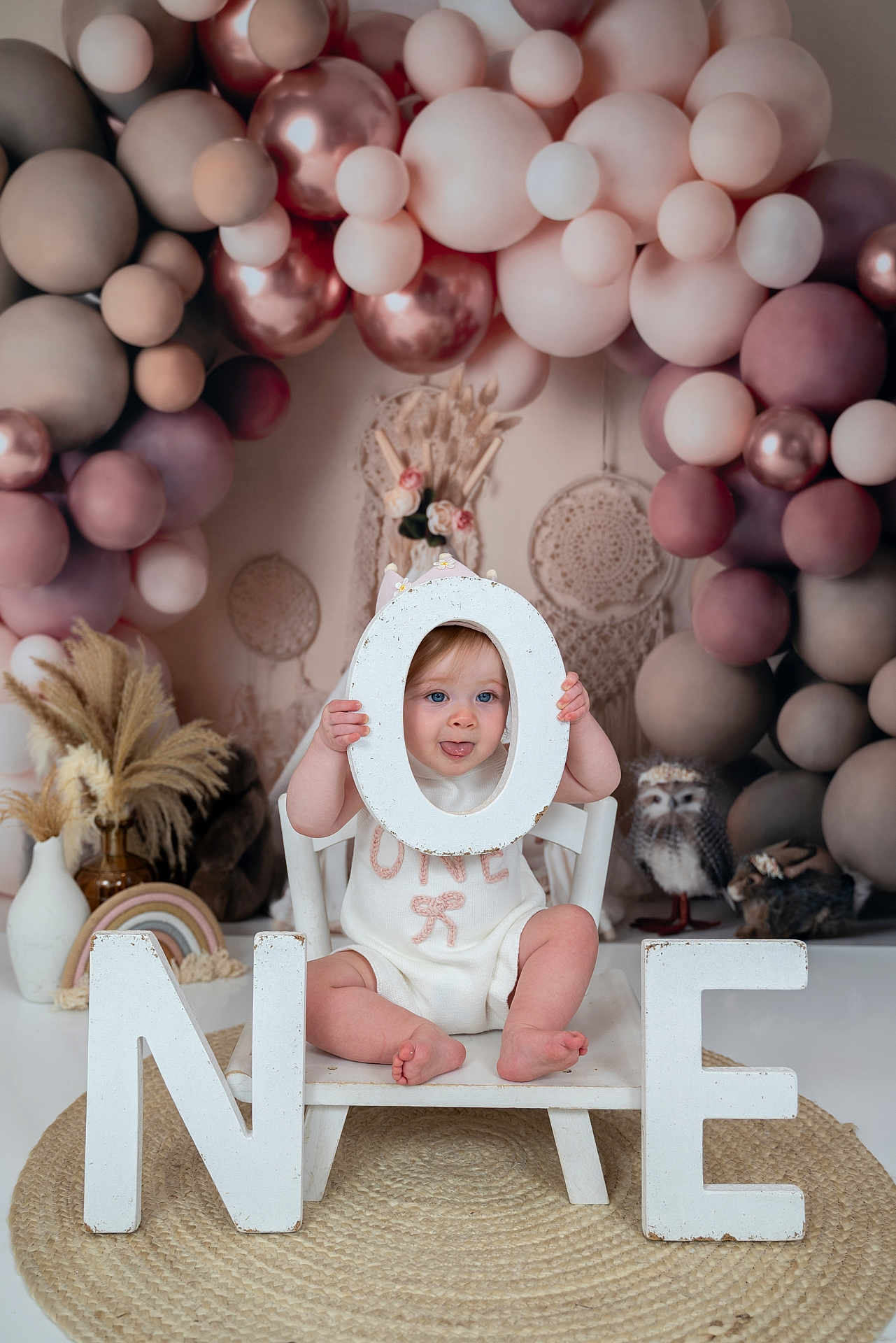 Orla is registered to the contest to win money with this photo: baby, child, one_year_old, birthday, balloons, decorations, letters, white_chair, playful, tongue_out, pastel_colors, boho_decor, round_rug, indoor, portrait, celebration, cute, small_feet, smiling, sitting