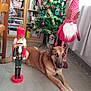 dog, christmas_tree, nutcracker, gnome, holiday_decor, indoor, floor, furniture, curtains, bookshelf, book, decoration, toy, brown_dog, festive, relaxed, christmas, pet, holiday, tile_floor