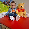 Joshua joined the competition — help win amazing prizes! cartoon_mural, casual_clothing, child, colorful, cute, happy, indoor, jeans, person, playroom, portrait, red_table, seated, smiling, sneakers, stuffed_animal, toddler, toy, winnie_the_pooh, young_child