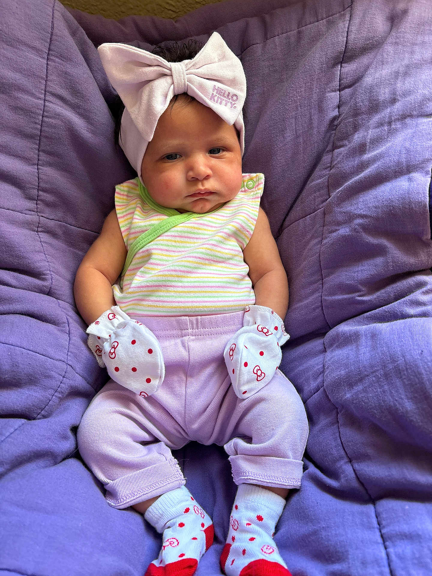 Kehlani is registered to the contest to win money with this photo: baby, infant, headband, hello_kitty, mittens, socks, purple_cushion, striped_clothing, pastel_colors, plush, seated, cute, soft_texture, casual, indoor, child, young_baby, portrait, comfortable, expression