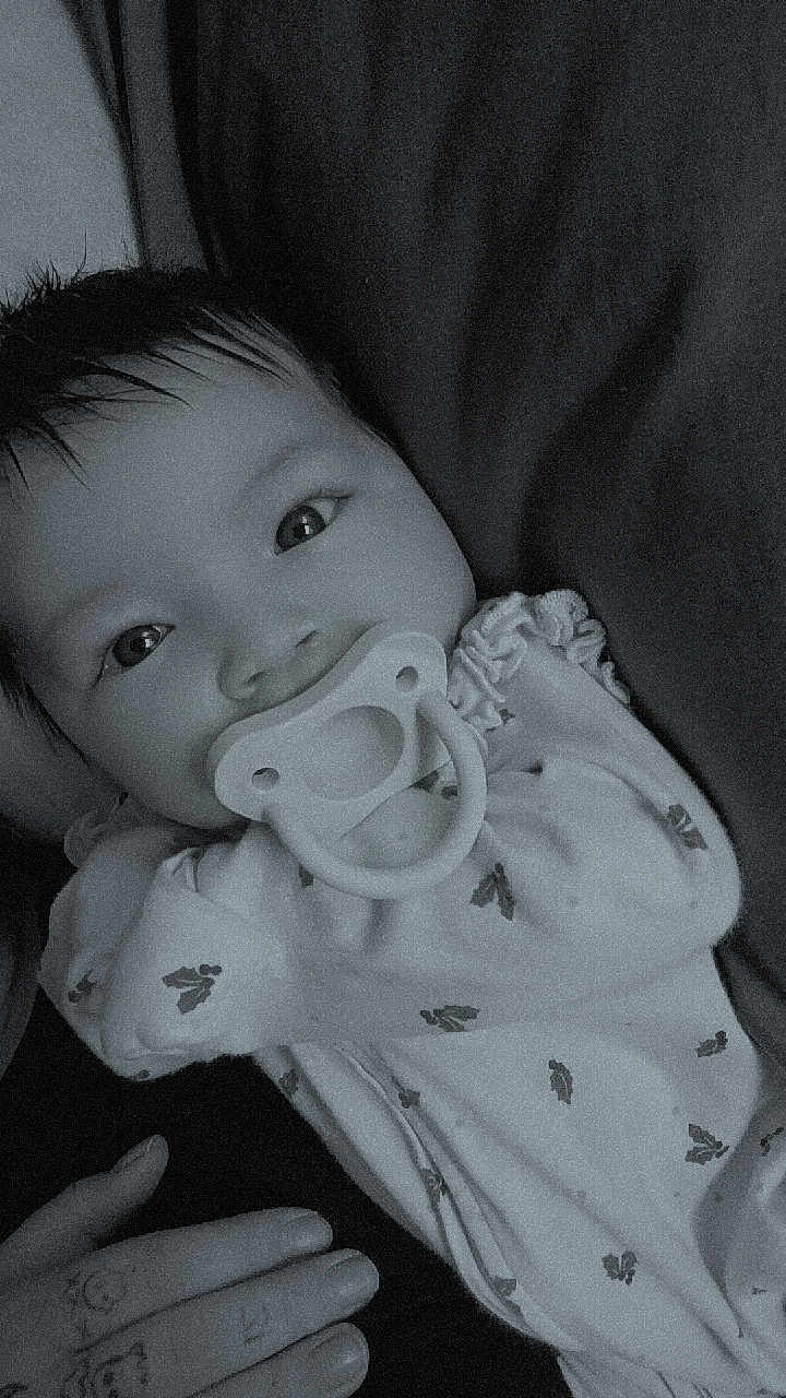 Freya is registered to the contest to win money with this photo: baby, infant, pacifier, pajamas, smile, eyes, hair, hand, tattoo, blanket, bed, lying_down, portrait, closeup, monochrome, black_and_white, cute, soft_texture, peaceful, nursery