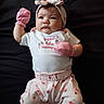 apparel, baby, baby_girl, bow, child, clothing, cute, dark_background, expression, face, headband, infant, lying_down, newborn, pants, person, pink_mittens, portrait, strawberry_pattern, white_shirt