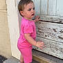 Maëna participe au concours pour gagner de l'argent avec cette photo : toddler, child, pink_clothing, short_sleeves, shorts, sneakers, earrings, concrete_step, wooden_door, peeling_paint, curious_expression, outdoor, side_view, standing, person, small_plant, wall, door_lock, daylight, casual_wear
