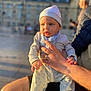 baby, child, infant, person, human_hand, clothing, hat, adult_hand, outdoor, sunlight, portrait, holding, warm_light, blurred_background, face, expression, casual, daytime, people, urban