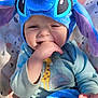 baby, child, blue_hat, stitch_hat, hand_in_mouth, light_blue_shirt, giraffe_print, elephant_print, blanket, patterned_background, cute, smiling, infant, portrait, indoors, soft_toys, toddler, face, clothing, happy