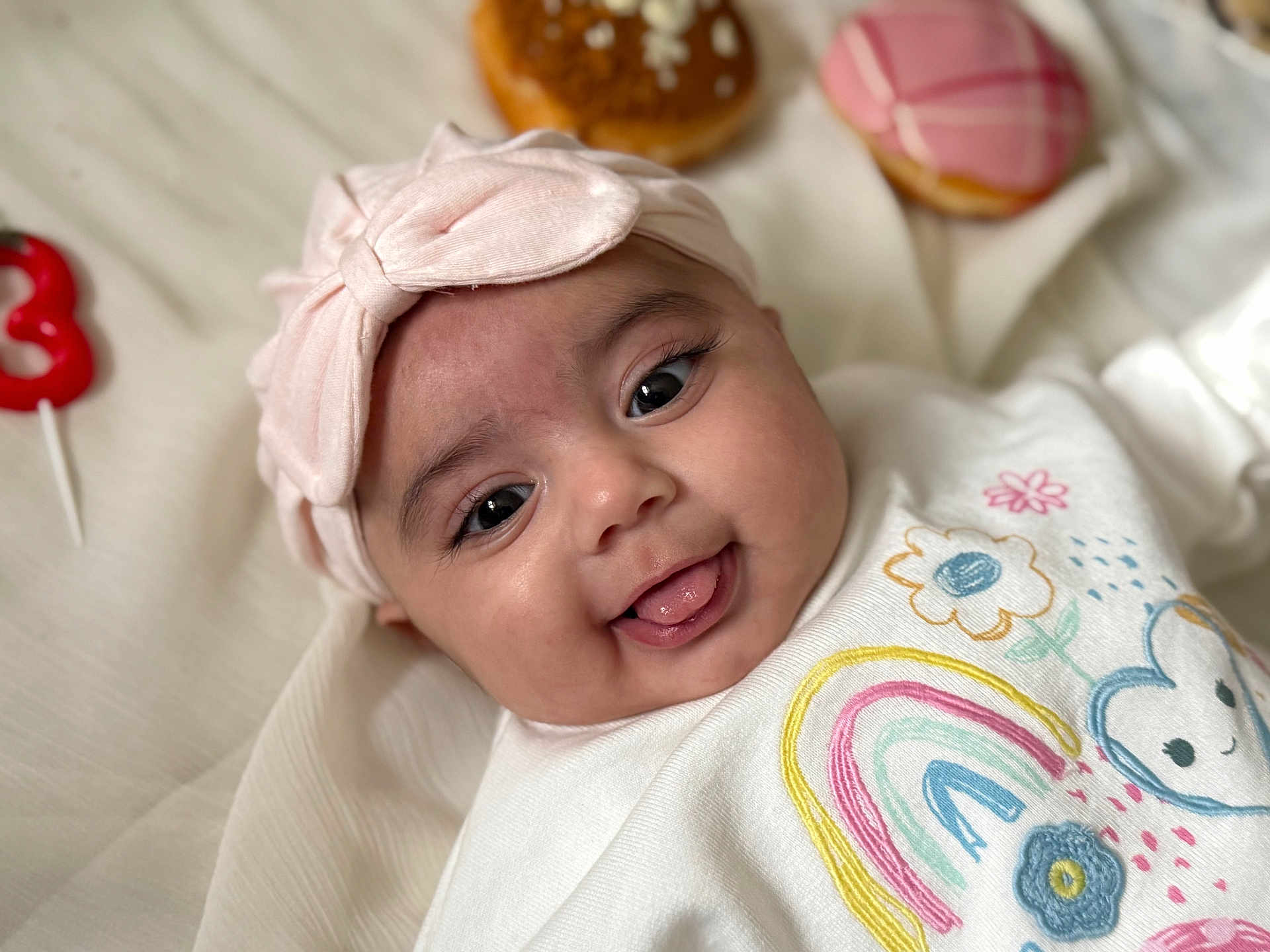Zariah joined the competition — help win amazing prizes! baby, infant, smiling, face, headband, bow, tongue, clothing, rainbow, flower, blanket, donut, toy, closeup, cute, happy, lying_down, indoors, soft_light, portrait