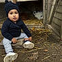 animal, black_hat, casual_clothing, child, curious_expression, fall, farmyard, ground, hat, leaves, nature, outdoor, pet, rabbit, sitting, stone, straw, toddler, white_shoes, wooden_hutch
