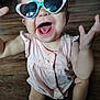 baby, child, sunglasses, heart_shaped, pink_dress, messy_face, laughing, happy, floor, wooden_floor, arms_raised, sitting, cute, playful, infant, indoor, clothing, person, portrait, smile