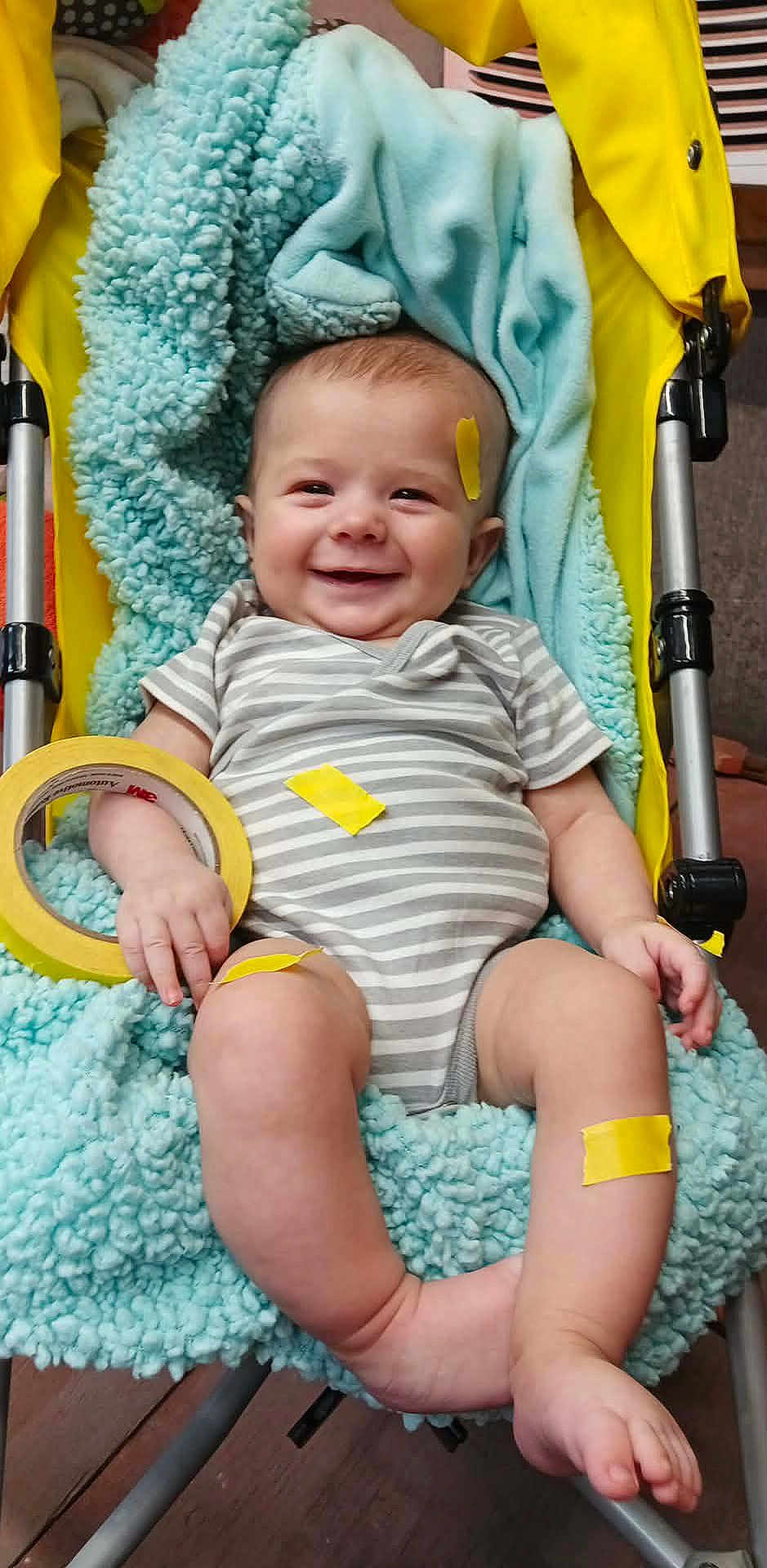Devin is registered to the contest to win money with this photo: baby, infant, stroller, blanket, tape, roll_of_tape, yellow_tape, blue_blanket, smiling, onesie, striped_clothing, legs, feet, hand, chubby_legs, indoor, cozy, seat, happy, portrait