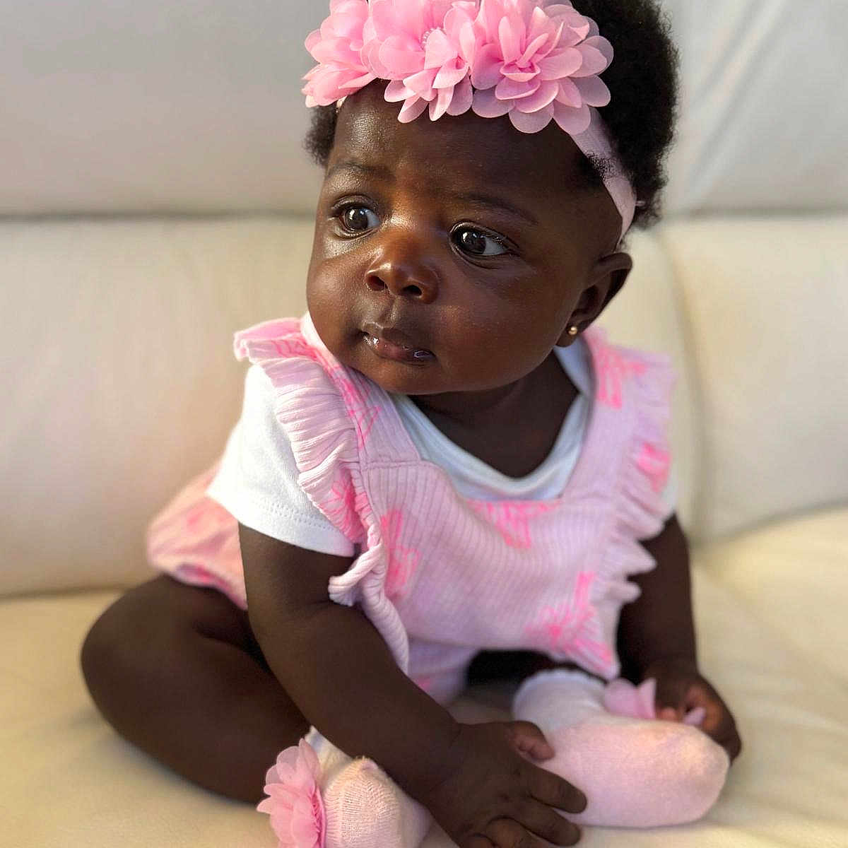 Anaya joined the competition — help win amazing prizes! baby, infant, child, girl, pink_headband, pink_outfit, sitting, curious, cute, portrait, indoors, couch, soft_lighting, flower_headband, sock, dark_skin, short_hair, earring, closeup, person