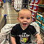 toddler, child, shopping_cart, store_aisle, toilet_paper, snack, lollipop, black_tshirt, messy_face, happy, grip, shopping, retail, grocery_store, baby_blanket, sitting, smiling, person, indoor, aisle
