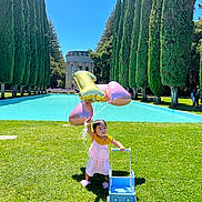 Maira is registered to the contest to win money with this photo: toddler, child, balloons, number_balloon, pink_dress, toy_stroller, grass, outdoor, sunny, trees, cypress_trees, reflecting_pool, blue_sky, shoes, play, summer, greenery, nature, happy, walking