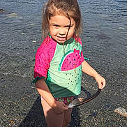 Scarlett is registered to the contest to win money with this photo: barefoot, beach, child, daytime, lake, mountains, nature, outdoor, person, playful, recreation, rocky_shore, smiling, stick, summer, sunlight, swim_shirt, water, watermelon_pattern, young_child