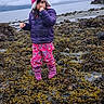 Sofie is registered to the contest to win money with this photo: beach, boots, child, cloudy_sky, coastal, cold_weather, colorful_clothing, curious, hat, jacket, nature, outdoor, pants, person, portrait, rocky_shore, seaweed, standing, thumb_sucking, water