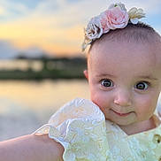 Minnie is registered to the contest to win money with this photo: baby, child, portrait, face, flower_headband, yellow_dress, cute, smiling, outdoor, sunset, nature, water, sky, blurry_background, happy, infant, person, closeup, arm, headwear