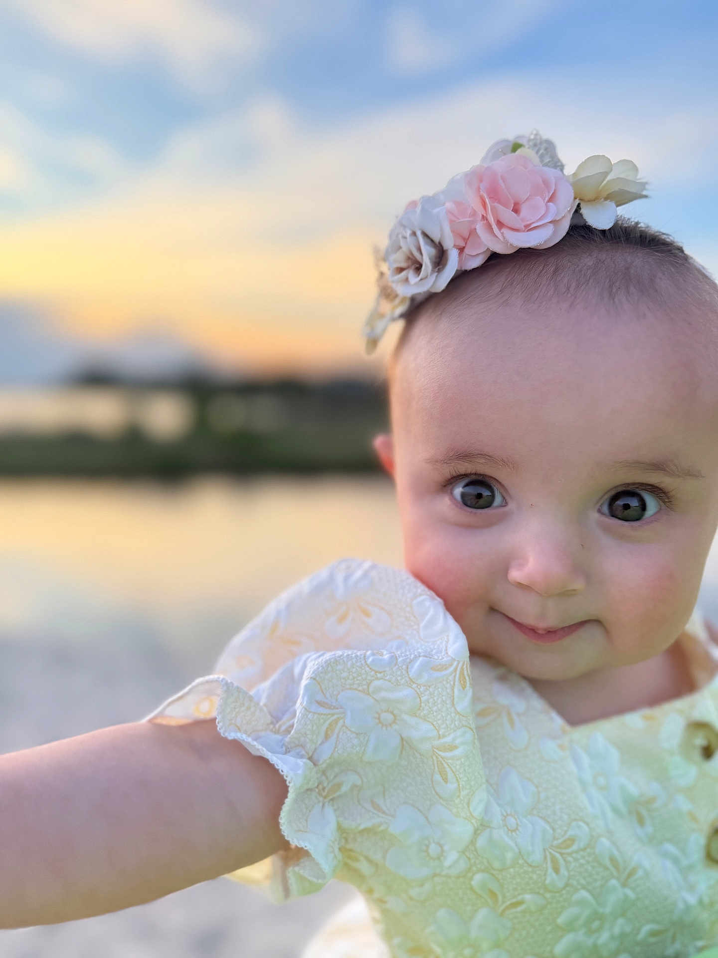 Minnie is registered to the contest to win money with this photo: baby, child, portrait, face, flower_headband, yellow_dress, cute, smiling, outdoor, sunset, nature, water, sky, blurry_background, happy, infant, person, closeup, arm, headwear