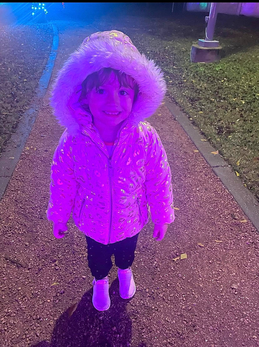 Holland is registered to the contest to win money with this photo: asphalt, electric_blue, freezing, fun, grass, happy, hood, joy, magenta, person, personal_protective_equipment, pink, plant, purple, road_surface, sleeve, smile, tints_and_shades, toddler, tree