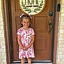 child, girl, smiling, dress, barefoot, door, welcome_mat, home_sign, hair_bows, outdoor, brick_wall, concrete, happy, person, front_door, plaid_bow, footwear, curly_hair, daylight, standing