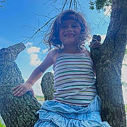 Holland is registered to the contest to win money with this photo: child, smiling, tree, outdoor, blue_sky, clouds, curly_hair, headband, striped_tank_top, blue_skirt, nature, branches, greenery, sunlight, happy, playful, summer, portrait, person, daytime