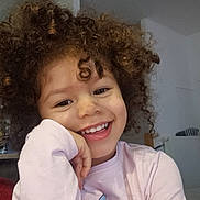 Maëlyss participe au concours pour gagner de l'argent avec cette photo : child, smiling, curly_hair, indoor, pink_shirt, happy, face, portrait, casual_clothing, resting_hand, young, cute, playful, natural_light, background, person, expression, closeup, head, joyful