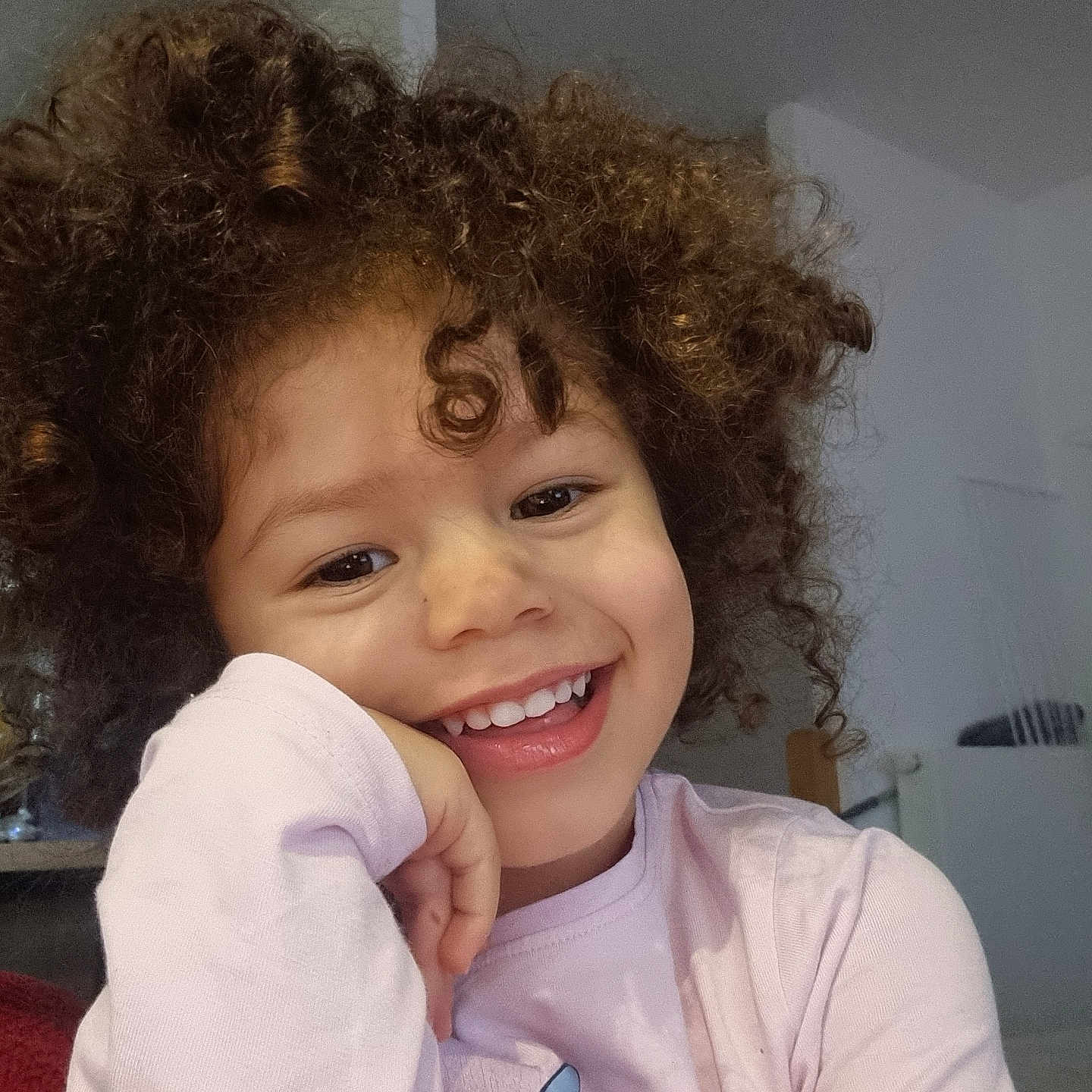 Maëlyss participe au concours pour gagner de l'argent avec cette photo : background, casual_clothing, child, closeup, curly_hair, cute, expression, face, happy, head, indoor, joyful, natural_light, person, pink_shirt, playful, portrait, resting_hand, smiling, young