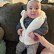 Maya is registered to the contest to win money with this photo: baby, child, infant, couch, fuzzy_vest, headband, bow, hand, person, clothing, shoes, pants, living_room, furniture, seated, cute, indoor, holding, smiling, portrait