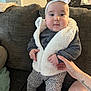 baby, child, infant, couch, fuzzy_vest, headband, bow, hand, person, clothing, shoes, pants, living_room, furniture, seated, cute, indoor, holding, smiling, portrait