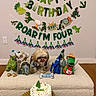 dog, birthday, cake, dinosaur, toys, party_hat, birthday_banner, fourth_birthday, indoor, celebration, festive, pet, shih_tzu, fluffy, carpet, wood_floor, cake_candles, plush_toys, birthday_bone, decorations