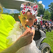 Kinsley joined the competition — help win amazing prizes! baby, child, headband, lollipop, hand, dress, white, outdoor, sunny, grass, golf_cart, people, trees, blue_sky, celebration, festive, person, summer, portrait, cute