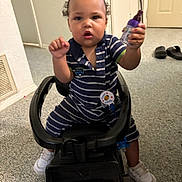 Amir is registered to the contest to win money with this photo: toddler, child, toy_car, indoor, striped_clothing, sneakers, car_seat, floor, door, slippers, hand, face, person, holding_object, curious_expression, hair, carpet, badge, pacifier, black_toy_car