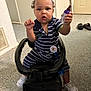 toddler, child, toy_car, indoor, striped_clothing, sneakers, car_seat, floor, door, slippers, hand, face, person, holding_object, curious_expression, hair, carpet, badge, pacifier, black_toy_car