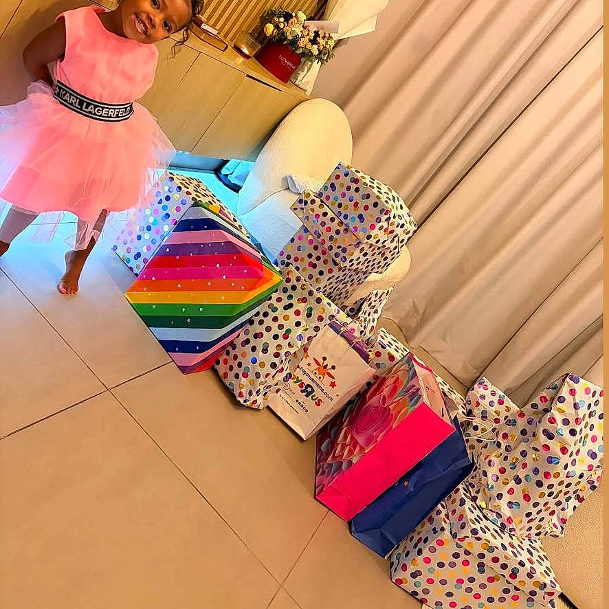 Anela is registered to the contest to win money with this photo: child, girl, smiling, pink_dress, indoor, gift_bags, presents, colorful, polka_dots, birthday, living_room, curtains, tile_floor, furniture, flowers, tissue_paper, portrait, standing, happy, social_media_screenshot