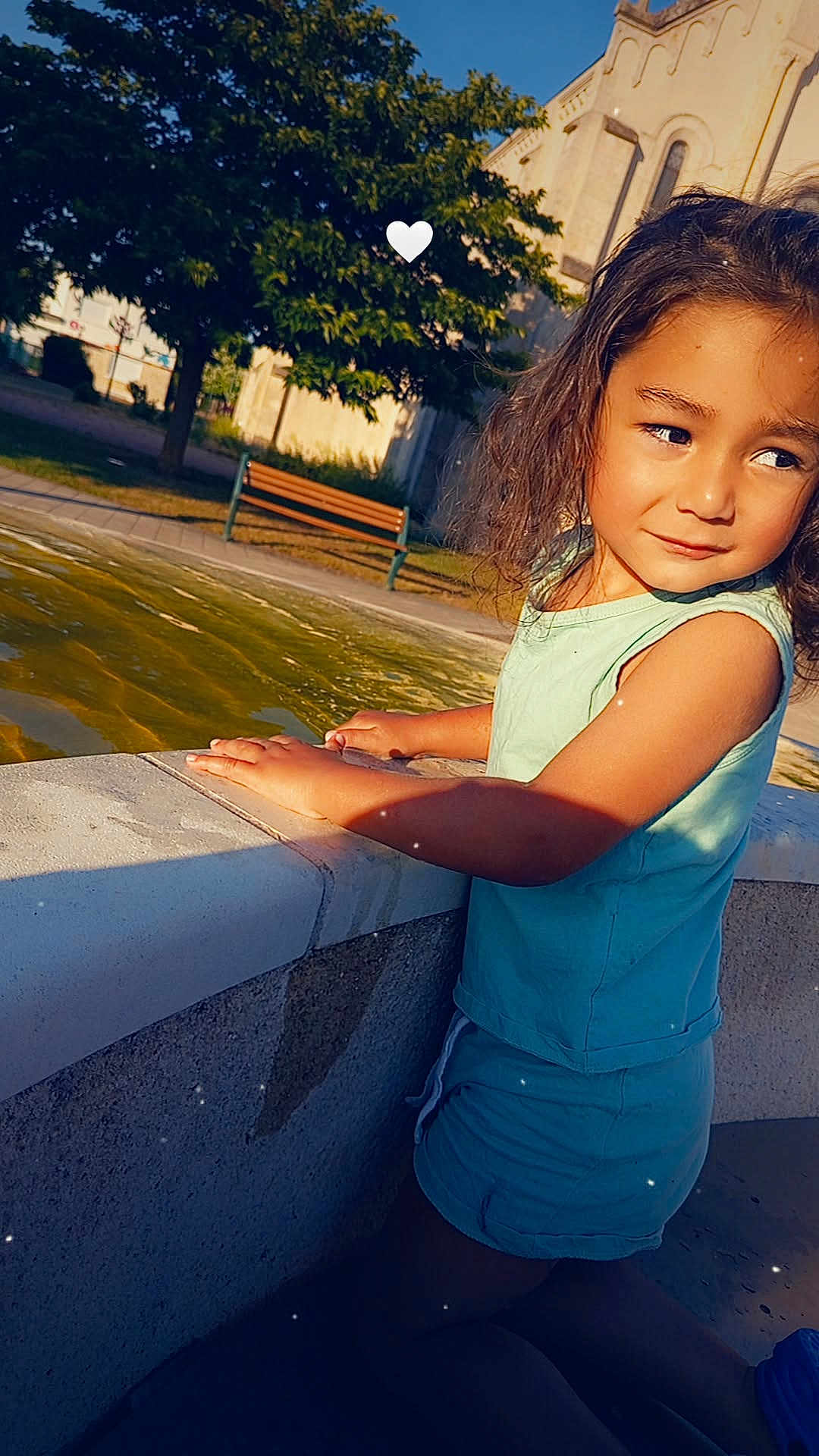 Anton participe au concours pour gagner de l'argent avec cette photo : child, curly_hair, water_fountain, outdoor, park, bench, tree, sunlight, shadow, summer_clothing, blue_shorts, blue_shirt, side_view, kneeling, thoughtful_expression, daytime, nature, skin, person, casual