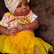 Amira joined the competition — help win amazing prizes! baby, child, tutu, yellow_skirt, headwrap, beige, brown_background, socks, pink_bows, colorful_top, cute, infant, lying_down, clothing, adorable, small_hands, portrait, indoors, fabric, fashion