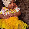 baby, child, tutu, yellow_skirt, headwrap, beige, brown_background, socks, pink_bows, colorful_top, cute, infant, lying_down, clothing, adorable, small_hands, portrait, indoors, fabric, fashion