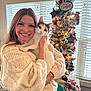 woman, cat, calico_cat, sweater, smiling, christmas_tree, holiday_decorations, window_blinds, bracelets, indoor, festive, happy, pet, holding, furniture, table, lights, ornaments, face, warm_lighting