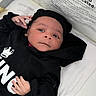 baby, infant, child, hoodie, black_clothing, lying_down, hand, face, eyes, expression, bed, blanket, warning_label, text, indoor, soft_lighting, cute, person, portrait, closeup