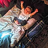 newborn, baby, infant, child, portrait, lying_down, smiling, face, arms_raised, clothing, hat, blanket, bed, pillow, indoors, home, text_on_clothing, soft_lighting, carpet, family