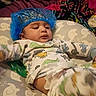 baby, baby_bedding, bed, blanket, blue_hat, casual_photo, child, clothing, dinosaur, face, home, indoors, infant, lying_down, nursery, onesie, pacifier, pillow, portrait, sleeping_area