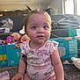 Maddie is registered to the contest to win money with this photo: baby, child, pigtails, sitting, carpet, toy, diaper_box, living_room, curious, indoor, clothing, pink_pants, patterned_shirt, plastic_bottle, couch, blanket, window, soft_light, face, person