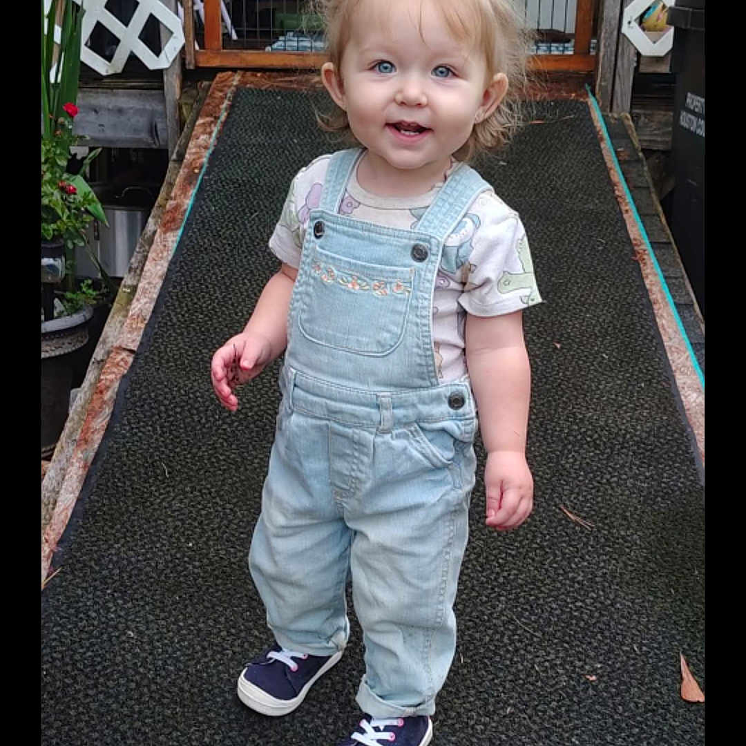 Everly Grace joined the competition — help win amazing prizes! bow, casual_clothing, child, daylight, denim, face, girl, hair, happy, lattice, outdoor, overalls, person, plants, ramp, smiling, sneakers, standing, toddler, wood