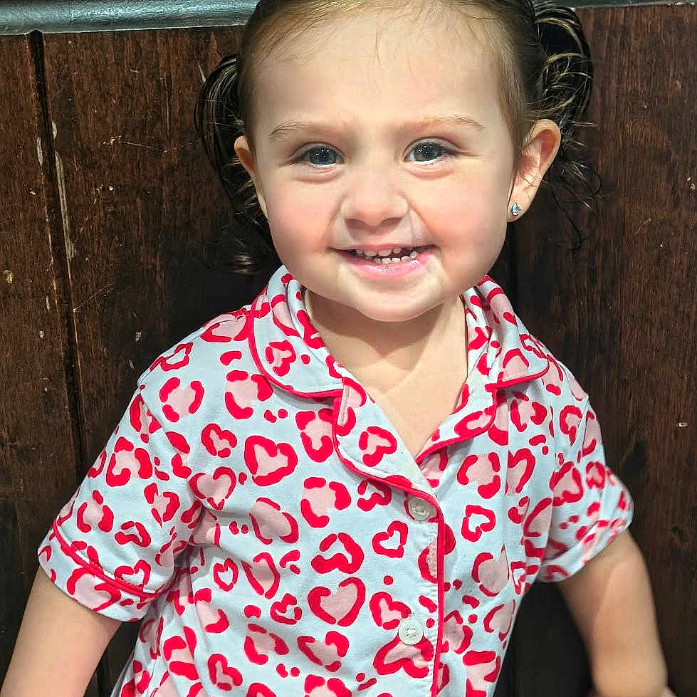 Layla joined the competition — help win amazing prizes! child, toddler, girl, smiling, portrait, pigtails, pajamas, heart_pattern, earrings, blue_eyes, indoors, wooden_background, seated, happy, face, person, closeup, casual_clothing, photography, adorable