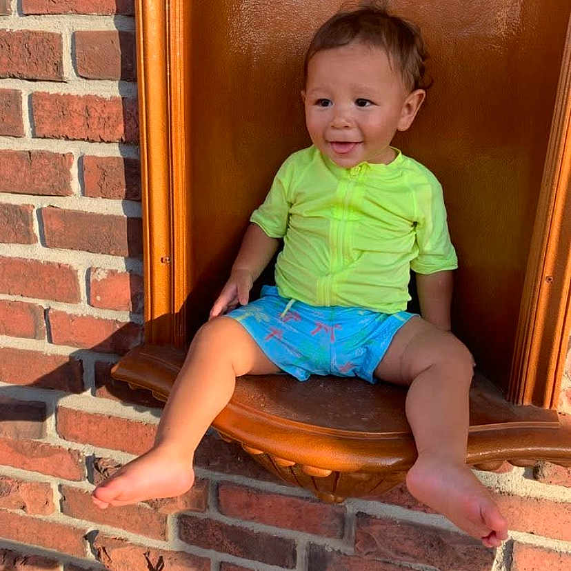 Asher joined the competition — help win amazing prizes! alcove, barefoot, brick_wall, casual_clothing, child, cute, foot, happy, head, neon_clothing, outdoor, person, portrait, shorts, sitting, skin, smiling, sunlight, toddler, young_child