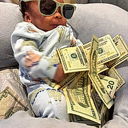 Nymir is registered to the contest to win money with this photo: baby, child, infant, sunglasses, money, dollar_bills, cash, portrait, sitting, couch, blanket, onesie, indoor, close_up, face, hair, fashion, funny, relaxed, photography