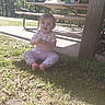 Willow is registered to the contest to win money with this photo: baby, child, toddler, sitting, outdoor, park, grass, picnic_area, picnic_table, shelter, trees, sunlight, casual_clothing, pink_clothing, long_sleeves, shoes, portrait, facial_expression, nature, daylight