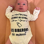 Elio a rejoint le concours — aidez-le/la à gagner de superbes lots ! baby, infant, onesie, clothing, text_on_clothing, face, arms, legs, padded_surface, curious_expression, wide_eyes, indoor, child, newborn, cute, person, laying_down, white_clothing, skin, human