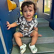 Mia joined the competition — help win amazing prizes! toddler, child, curly_hair, black_and_white_clothing, crocs, playground, steps, outdoor, daylight, grass, fence, house, blue_play_structure, smiling, sitting, cute, young_child, casual_clothing, shoes, person