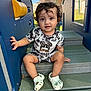 toddler, child, curly_hair, black_and_white_clothing, crocs, playground, steps, outdoor, daylight, grass, fence, house, blue_play_structure, smiling, sitting, cute, young_child, casual_clothing, shoes, person